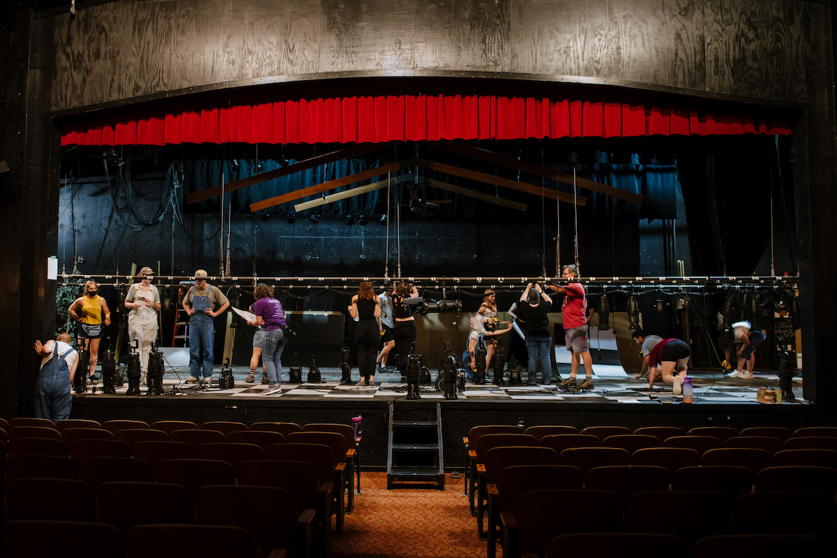 Several stage technicians work on a lighting rig while standing on a production stage.
