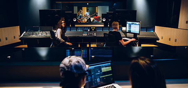 In the background, three guitarists are seen in a recording booth. In the mid-ground in focus are two Full Sail artists sitting at a mixing board. In the foreground and out of focus are two more artists sitting at a laptop