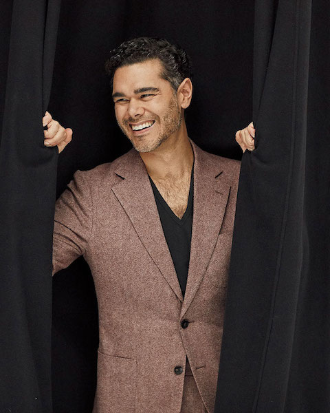 Press photo of Elliot Madore smiling, opening black curtains and wearing a brown suit