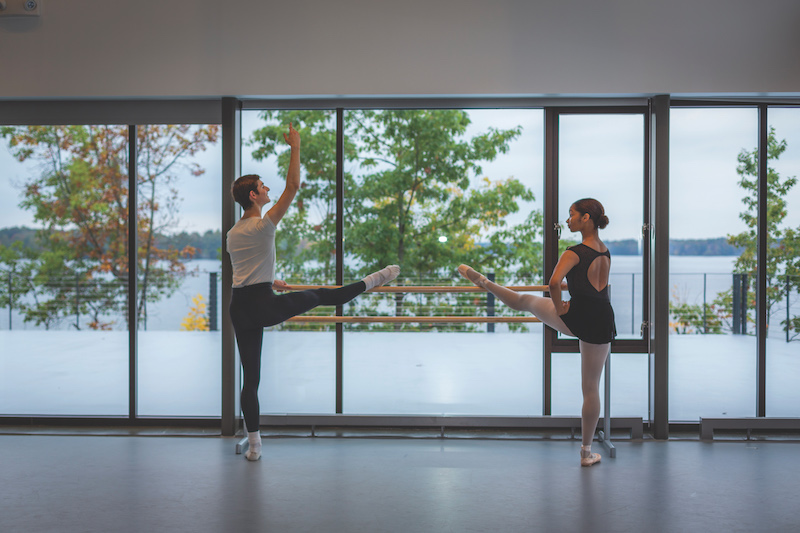 Two students in a dance studio face each other with one leg pointed on a ballet barre, the windows behind them looking out on trees and a lake