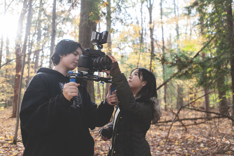 Two students stand in the woods, one holding a camera rig and the student at the right adjusting the camera