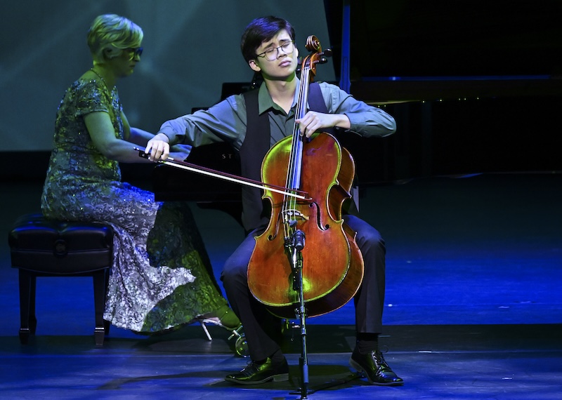 A cellist sits with his eyes closed, with his accompanist in the background