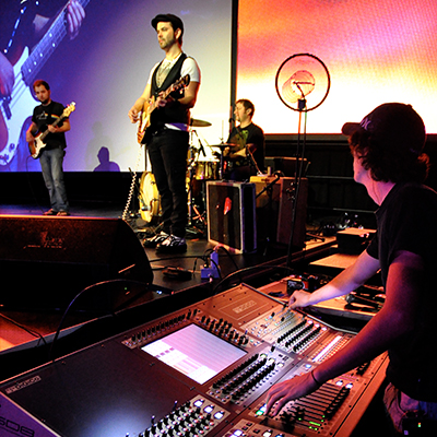 Full Sail student mixing at a board while looking at two guitar players and a drummer on stage with a large projection screen in the background