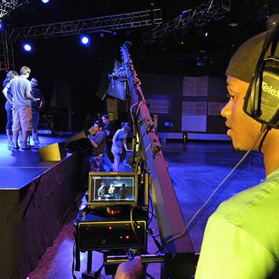 Full Sail student in foreground wearing headphones and filming three students on stage while students in the background set up speakers