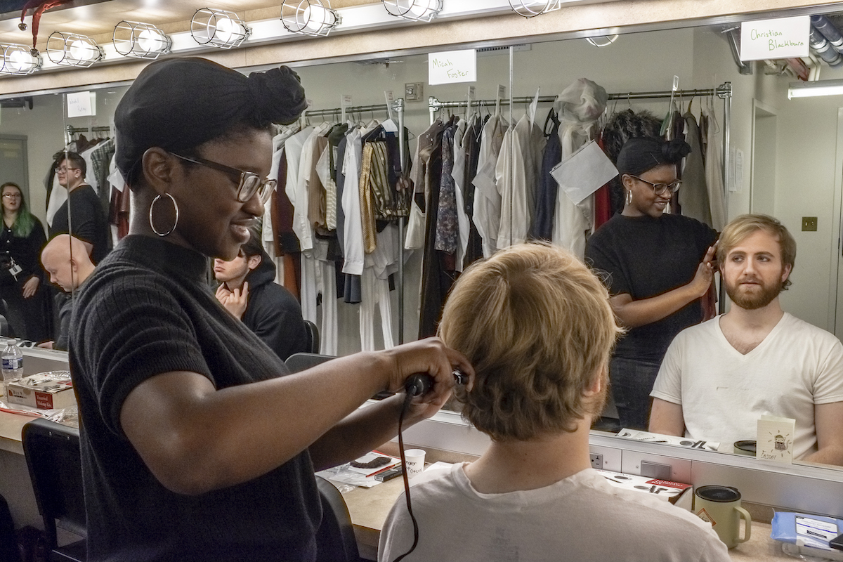 UNCG backstage photo of Falstaff. A hairdresser curls the hair of an actor in her chair as they face the dressing room mirrors. Other actors interact and several costumes are hung in the background.